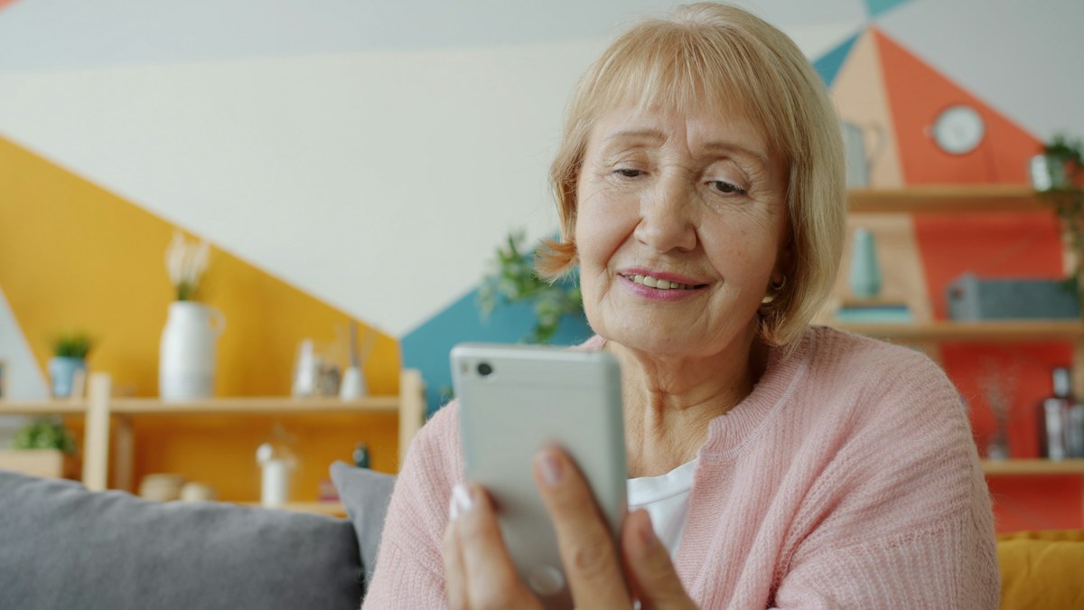 Starsza kobieta w różowym swetrze uśmiecha się, patrząc w ekran telefonu