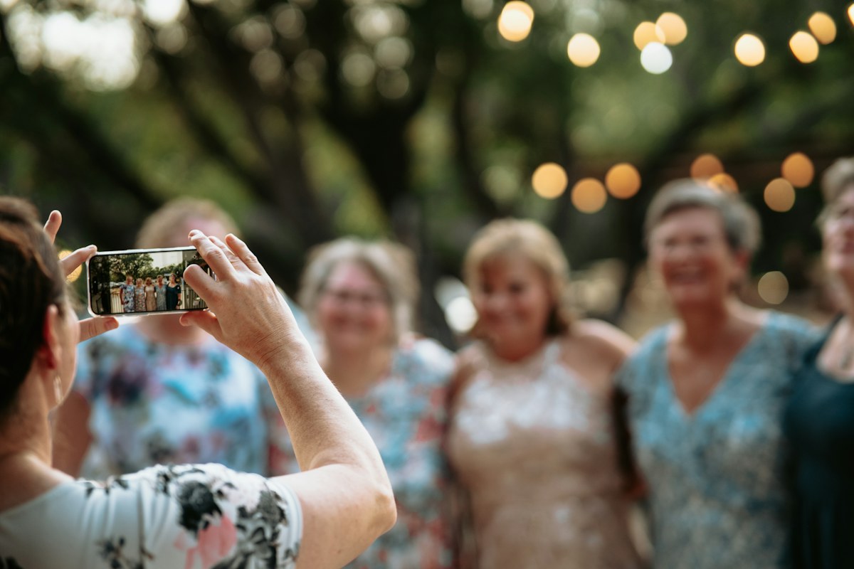 Gość weselny fotografuje telefonem grupę uśmiechniętych kobiet pod girlandą lampek